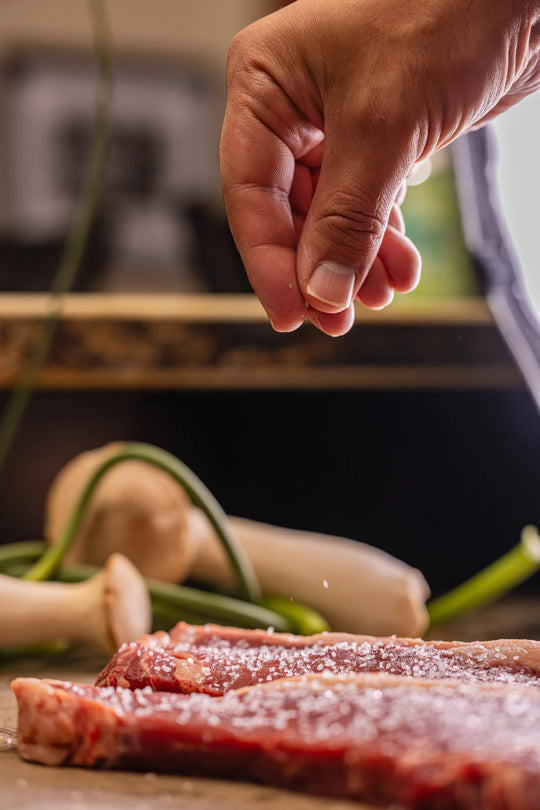 Hand seasoning raw meat with salt on a wooden cutting board.
